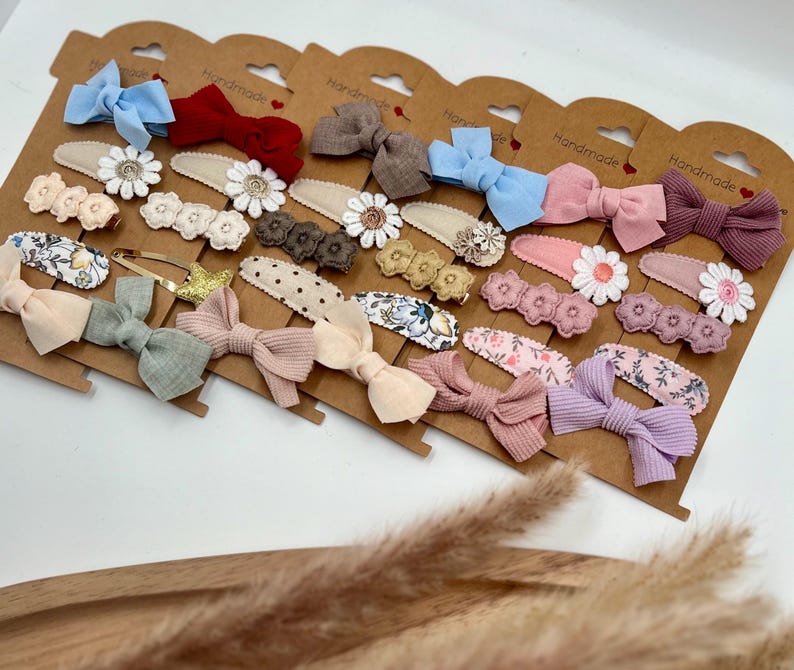 Puede incluir: Un conjunto de 18 pinzas para el pelo hechas a mano con una variedad de colores y patrones. Las pinzas incluyen lazos, flores y lunares. Las pinzas se muestran en una tarjeta marr&oacute;n con la palabra "Handmade" impresa en cada tarjeta.