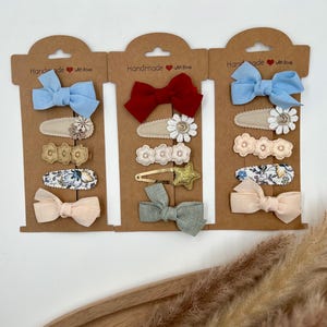 Puede incluir: Tres juegos de pinzas para el pelo en cartulina marr&oacute;n con el texto "Handmade with love". Cada juego incluye un lazo, un dise&ntilde;o floral y una pinza de tela estampada. Los lazos son de color azul, rojo y rosa claro.