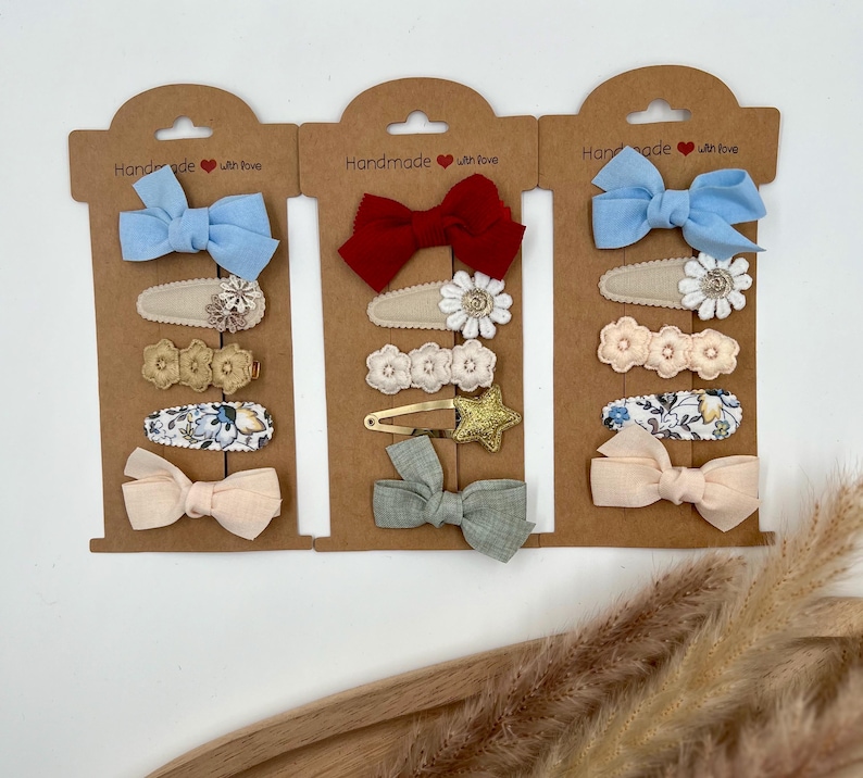Puede incluir: Tres juegos de pinzas para el pelo en cartulina marr&oacute;n con el texto "Handmade with love". Cada juego incluye un lazo, un dise&ntilde;o floral y una pinza de tela estampada. Los lazos son de color azul, rojo y rosa claro.