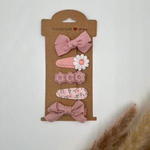 Puede incluir: Conjunto de cinco pinzas para el pelo de color rosa con dise&ntilde;os florales y de lazo. Las pinzas se muestran en una tarjeta marr&oacute;n con el texto "Handmade with love".