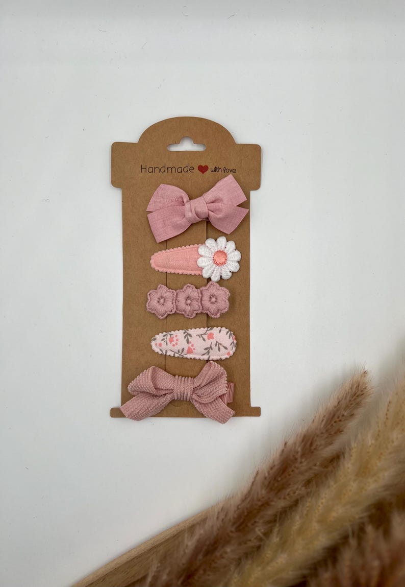 Puede incluir: Conjunto de cinco pinzas para el pelo de color rosa con dise&ntilde;os florales y de lazo. Las pinzas se muestran en una tarjeta marr&oacute;n con el texto "Handmade with love".