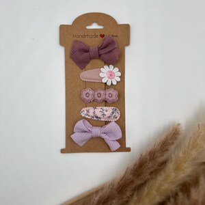 Puede incluir: Conjunto de cinco pinzas para el pelo en una tarjeta marr&oacute;n con el texto "Handmade with love". Las pinzas son moradas y rosas con dise&ntilde;os florales y de lazo.