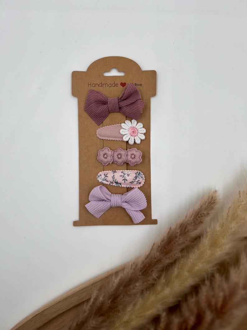 Puede incluir: Conjunto de cinco pinzas para el pelo en una tarjeta marr&oacute;n con el texto "Handmade with love". Las pinzas son moradas y rosas con dise&ntilde;os florales y de lazo.