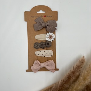 Puede incluir: Conjunto de cinco pinzas para el pelo en una tarjeta marr&oacute;n con el texto "Handmade with love". El conjunto incluye dos lazos marrones, dos pinzas florales con flores marrones y blancas, y una pinza de lunares.
