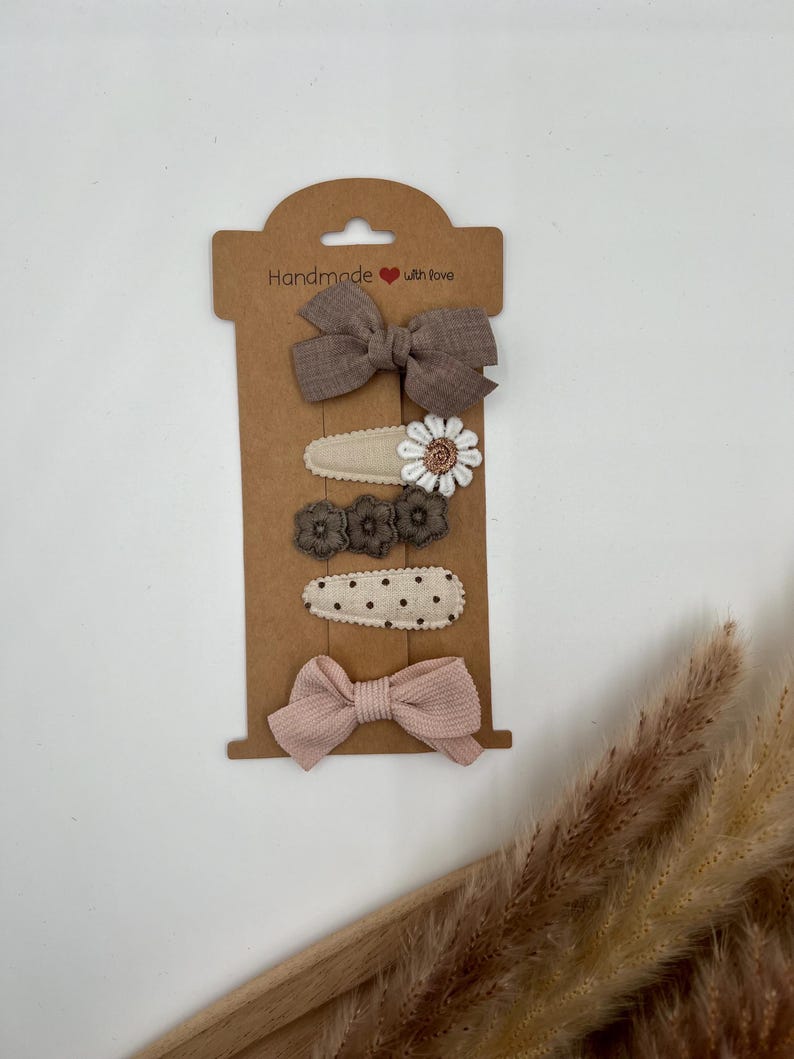 Puede incluir: Conjunto de cinco pinzas para el pelo en una tarjeta marr&oacute;n con el texto "Handmade with love". El conjunto incluye dos lazos marrones, dos pinzas florales con flores marrones y blancas, y una pinza de lunares.
