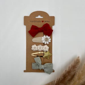 Puede incluir: Conjunto de cinco pinzas para el pelo en una tarjeta marr&oacute;n con el texto "Handmade with love". Las pinzas incluyen un lazo rojo, una flor blanca, una estrella dorada y dos lazos verdes.