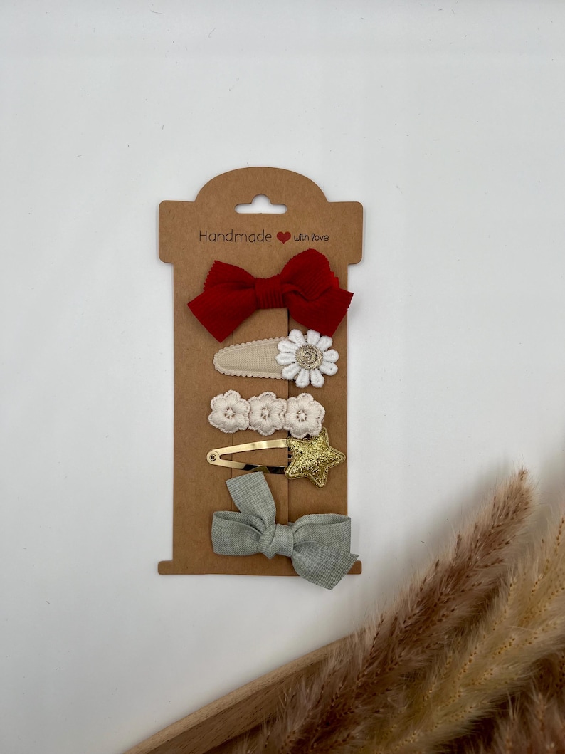 Puede incluir: Conjunto de cinco pinzas para el pelo en una tarjeta marr&oacute;n con el texto "Handmade with love". Las pinzas incluyen un lazo rojo, una flor blanca, una estrella dorada y dos lazos verdes.