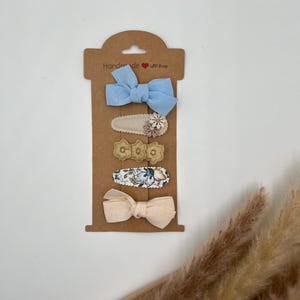 Puede incluir: Conjunto de cinco pinzas para el pelo en una tarjeta marr&oacute;n con el texto "Handmade with love". El conjunto incluye dos lazos de tela, uno azul claro y otro rosa claro, y tres pinzas con estampado floral.