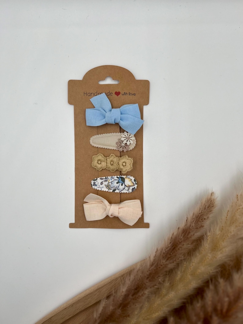 Puede incluir: Conjunto de cinco pinzas para el pelo en una tarjeta marr&oacute;n con el texto "Handmade with love". El conjunto incluye dos lazos de tela, uno azul claro y otro rosa claro, y tres pinzas con estampado floral.