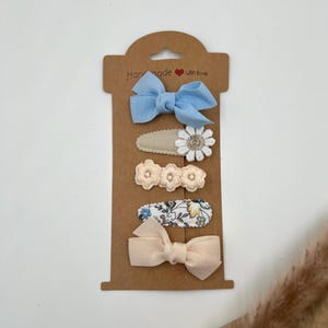 Puede incluir: Conjunto de cinco pinzas para el pelo en una tarjeta marr&oacute;n con el texto "Handmade with love". Las pinzas presentan un lazo azul, una flor blanca, dos flores de color melocot&oacute;n y un estampado floral.