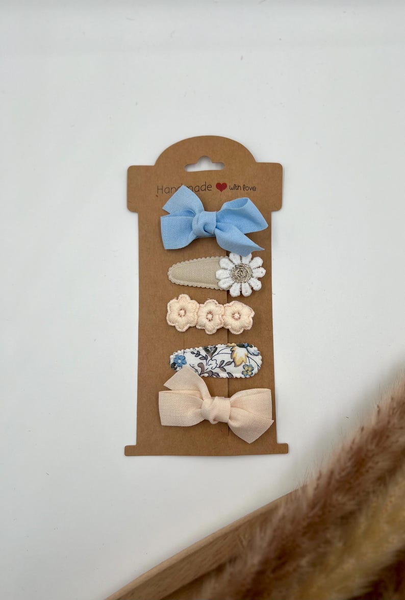 Puede incluir: Conjunto de cinco pinzas para el pelo en una tarjeta marr&oacute;n con el texto "Handmade with love". Las pinzas presentan un lazo azul, una flor blanca, dos flores de color melocot&oacute;n y un estampado floral.