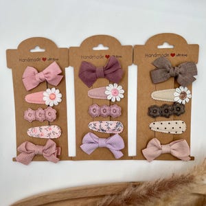 Puede incluir: Tres juegos de pinzas para el pelo en cartulina marr&oacute;n. Cada juego incluye un lazo, una pinza floral y una pinza estampada. Los lazos son en tonos rosa, morado y marr&oacute;n. Las pinzas florales tienen flores blancas con centros rosas. Las pinzas estampadas tienen estampados florales rosas, lunares marrones y estampados florales rosas. La cartulina tiene el texto "Handmade with love" en cada juego.