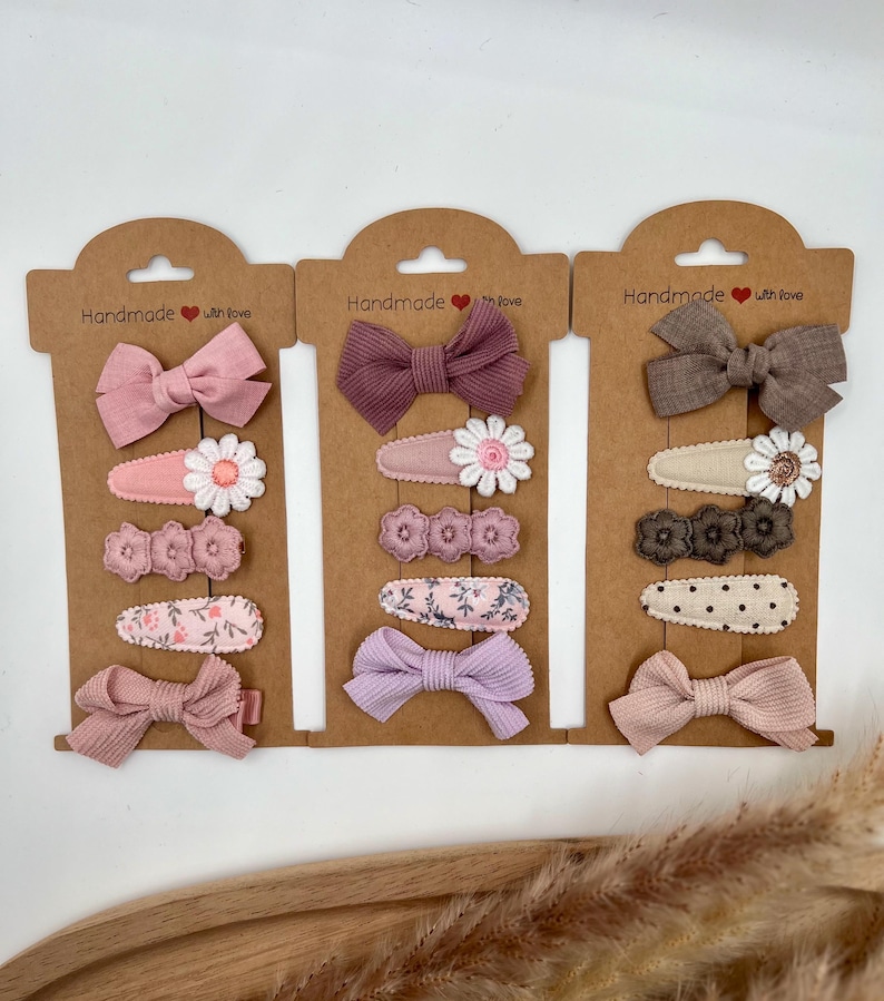 Puede incluir: Tres juegos de pinzas para el pelo en cartulina marr&oacute;n. Cada juego incluye un lazo, una pinza floral y una pinza estampada. Los lazos son en tonos rosa, morado y marr&oacute;n. Las pinzas florales tienen flores blancas con centros rosas. Las pinzas estampadas tienen estampados florales rosas, lunares marrones y estampados florales rosas. La cartulina tiene el texto "Handmade with love" en cada juego.