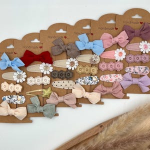 Puede incluir: Surtido de pinzas para el pelo hechas a mano con varios colores y patrones, incluyendo flores, lunares y colores s&oacute;lidos. Las pinzas se muestran en tarjetas marrones con la palabra "Handmade" impresa en ellas.