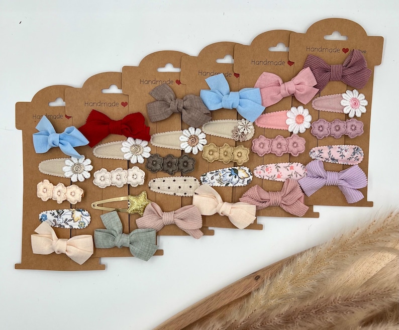 Puede incluir: Surtido de pinzas para el pelo hechas a mano con varios colores y patrones, incluyendo flores, lunares y colores s&oacute;lidos. Las pinzas se muestran en tarjetas marrones con la palabra "Handmade" impresa en ellas.
