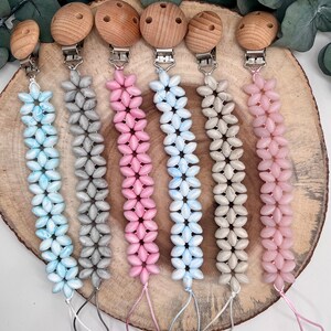 Könnte beinhalten: Sechs Holz-Beißringe mit unterschiedlich farbigen Perlen. Die Beißringe sind an einem Clip befestigt und haben eine Schlaufe am Ende. Die Farben der Perlen sind blau, grau, rosa, hellblau, beige und rosa.