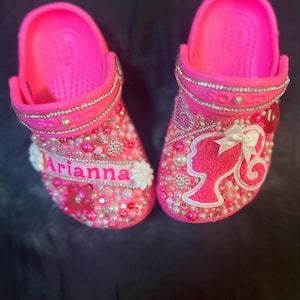 Pode incluir: Sandálias rosa personalizadas para crianças com uma silhueta de Barbie, strass e pérolas. As sandálias são decoradas com o nome "Arianna".