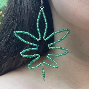 May include: A pair of green beaded earrings in the shape of a leaf. The earrings are made with small, iridescent green beads strung on wire, creating a delicate, open design. The earrings hang from silver-colored ear wires.