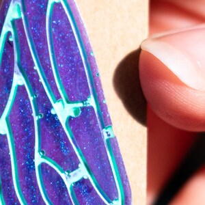 May include: A purple and blue resin pendant with a glittery finish and a white and green design. The pendant is shaped like a wing.