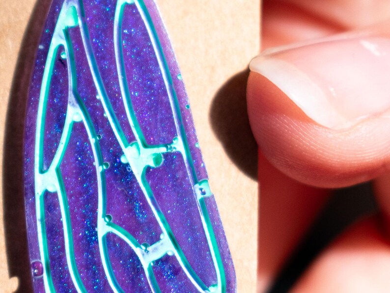 May include: A purple and blue resin pendant with a glittery finish and a white and green design. The pendant is shaped like a wing.