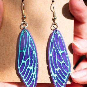 May include: A pair of iridescent purple and green earrings shaped like cicada wings. The earrings have a silver hook and are made of a translucent material.