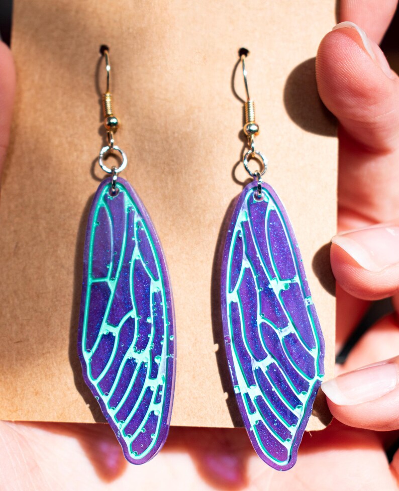 May include: A pair of iridescent purple and green earrings shaped like cicada wings. The earrings have a silver hook and are made of a translucent material.