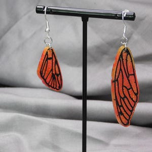 May include: A pair of earrings featuring orange and yellow butterfly wings with black outlines. The wings are translucent and have a glittery finish.