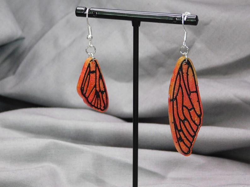 May include: A pair of earrings featuring orange and yellow butterfly wings with black outlines. The wings are translucent and have a glittery finish.