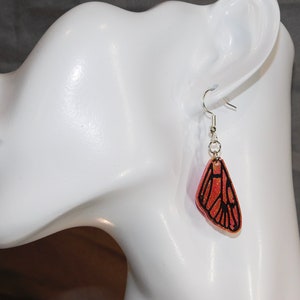 May include: A pair of dangle earrings featuring a butterfly wing design. The wings are made of resin and have a glittery, orange finish with black outlines. The earrings are attached to silver hooks.