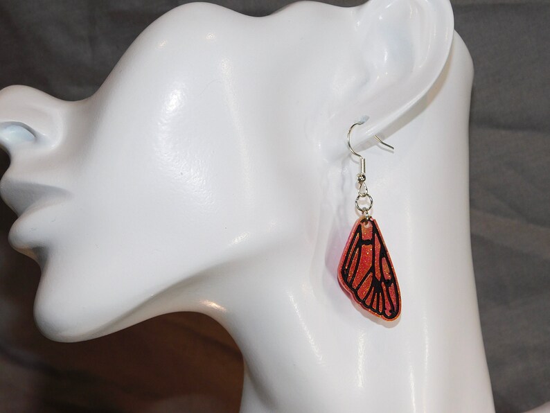 May include: A pair of dangle earrings featuring a butterfly wing design. The wings are made of resin and have a glittery, orange finish with black outlines. The earrings are attached to silver hooks.