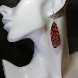 May include: A pair of dangle earrings featuring a red and gold cicada wing design. The wings are outlined in black and have a glittery finish.
