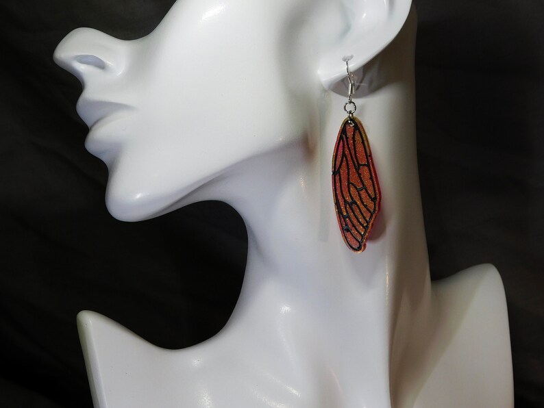 May include: A pair of dangle earrings featuring a red and gold cicada wing design. The wings are outlined in black and have a glittery finish.
