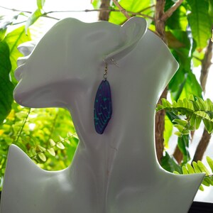 May include: A pair of earrings featuring a blue and green iridescent butterfly wing design. The earrings are dangling and have a gold hook.