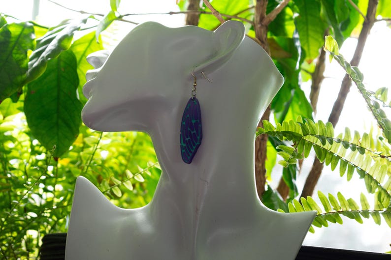May include: A pair of earrings featuring a blue and green iridescent butterfly wing design. The earrings are dangling and have a gold hook.