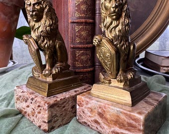 Puede incluir: Un par de sujetalibros antiguos con forma de león dorado, cada uno sosteniendo un escudo, sobre bases rectangulares de mármol. Los sujetalibros se exhiben con libros antiguos encuadernados en cuero, creando un tema decorativo clásico.