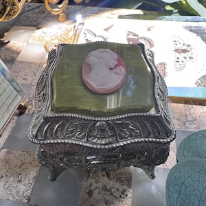 May include: A square jewelry box with an ornate silver-toned metal base featuring intricate relief patterns and four small feet. The lid is topped with a smooth, olive-green square panel centered with a pink and white oval cameo portrait.