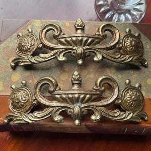 May include: Two matching brass drawer pulls featuring an ornate urn-shaped center, scrolling leaf motifs, and circular floral rosettes on each end. The metal hardware has a weathered, aged patina with visible dark highlights in the recessed areas.