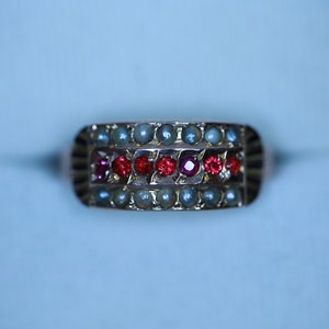 May include: A gold ring featuring a row of red gemstones surrounded by two rows of white pearls.