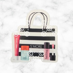 以下が含まれることがあります： 白と黒のストライプのショッピングバッグの形をしたステッカーで、美容製品が入っています。「Summer Campfires」と書かれた赤いチューブ、青とオレンジの容器、そして「Sailing」と書かれたピンクのリップグロスが含まれています。