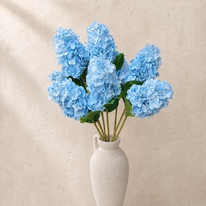 May include: A bouquet of light blue hydrangea flowers with green leaves in a cream-colored ceramic vase. The flowers have a full, rounded shape and are arranged in a visually appealing display. The vase has a textured surface and a handle.