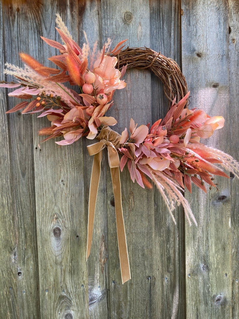 May include: A fall-themed wreath with a brown grapevine base. The wreath is decorated with pink and orange dried flowers, brown velvet ribbon, and beige pampas grass.