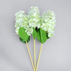 May include: Three artificial hydrangea stems with light green flower clusters and large green leaves. The stems are a light yellow color. The flowers are a soft, muted green and white. The background is a solid light gray.