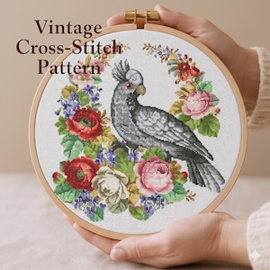 May include: A cross-stitch pattern featuring a gray bird with a crest, surrounded by a floral wreath of red, pink, and purple flowers. The text "Vintage Cross-Stitch Pattern" is at the top. The embroidery is held in a wooden hoop.