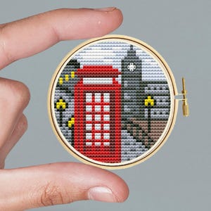 Può includere: Un telaio da ricamo a punto croce con una cabina telefonica rossa e un paesaggio urbano. Il disegno comprende una torre dell'orologio e altri edifici, con lampioni gialli. Il ricamo è in un telaio di legno.