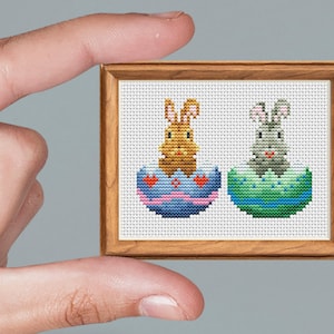 May include: A framed cross-stitch featuring two Easter bunnies emerging from decorated eggs. One bunny is tan, the other gray. The eggs are blue and green, with heart and stripe patterns. The frame is wood.