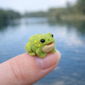 May include: A small, handmade green frog figurine with yellow spots and black eyes. The frog is made of a fuzzy material and has a smiling expression. The frog is resting on a finger, with a blurred water background.