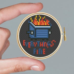 May include: A cross-stitch embroidery hoop with a black background. The design features a blue skip with orange flames and the text "Everything's Fine" in red and blue. The hoop is held by a person.