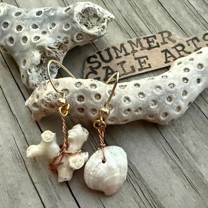May include: A pair of dangle earrings featuring gold-toned wire hooks and accents. One earring displays a small, textured white coral branch, while the other features a smooth, off-white seashell. The earrings are displayed near a sign reading SUMMER SALE ARTS.