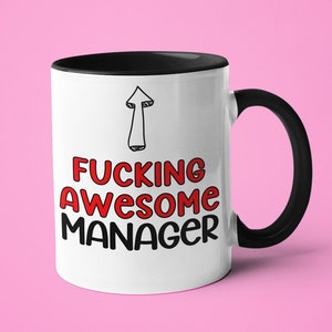 May include: White ceramic mug with a black handle and rim. The mug features the text "FUCKING AWESOME MANAGER" in red and black, with an upward-pointing arrow graphic. The mug is set against a pink background.