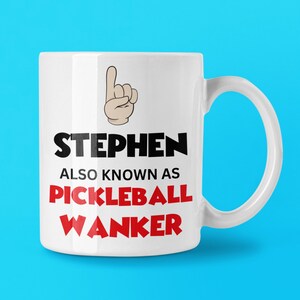 May include: White ceramic mug with the name "STEPHEN" in black, and "ALSO KNOWN AS PICKLEBALL WANKER" in red. A cartoon hand with the index finger pointing up is above the name. The mug has a white handle.